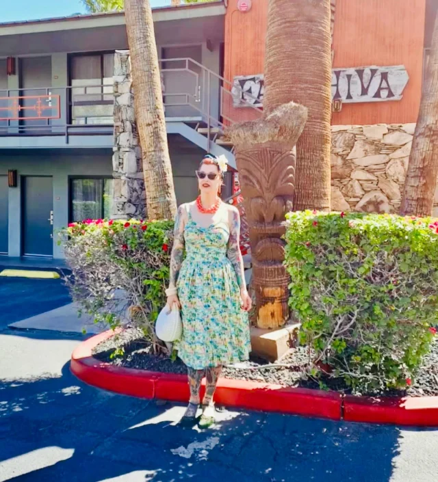 A woman stands in front of a building with a tiki statue and palm trees.