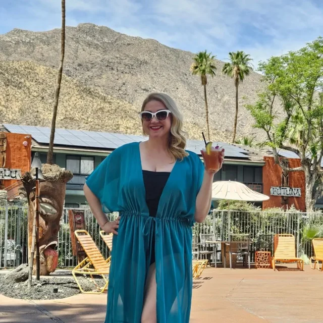 A woman in a teal dress holding a drink in front of a resort pool area with a mountain view.