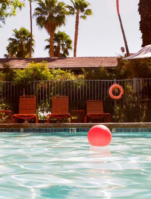 A swimming pool with a red ball floating in it, surrounded by orange chairs and a metal fence, with palm trees in the background.
